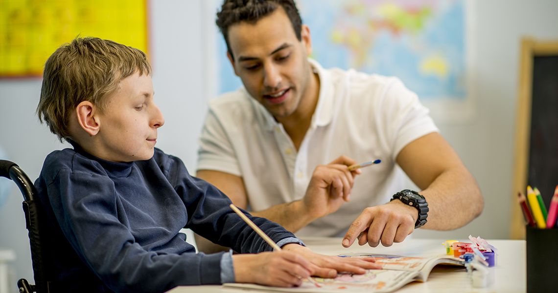 Boy in a wheelchair holding a pencil writing with a man helping him.