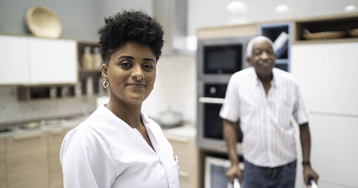 Woman in a white healthcare worker uniform, looking proudly at the camera, with an elderly man using a walker in the background