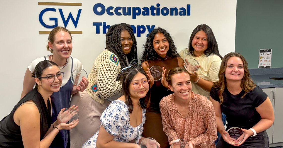 8 Occupational Therapy Students holding circle awards in front of GW OT sign