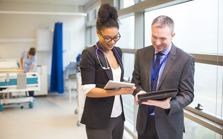 Female clinician with a stethoscope around her neck looking down at a notebook standing next to a man in a suit doing the same