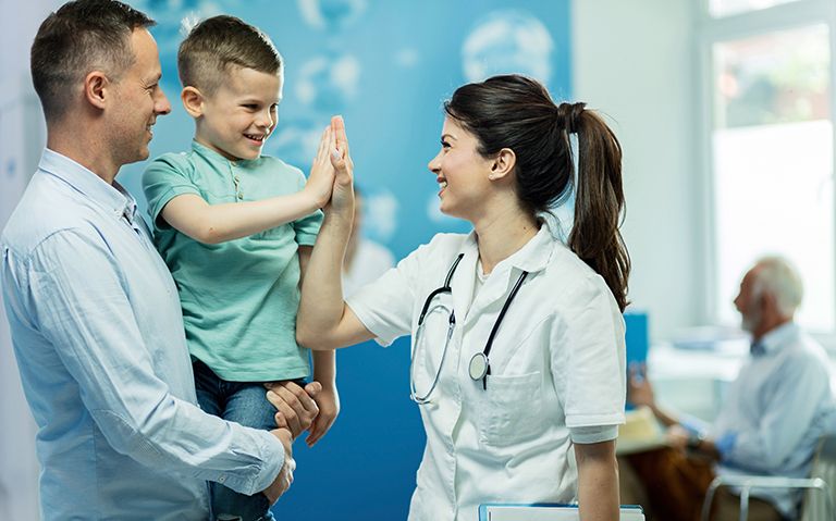Female doctor wearing stethoscope around her neck smiling and giving a high five to a child who is being held by his dad