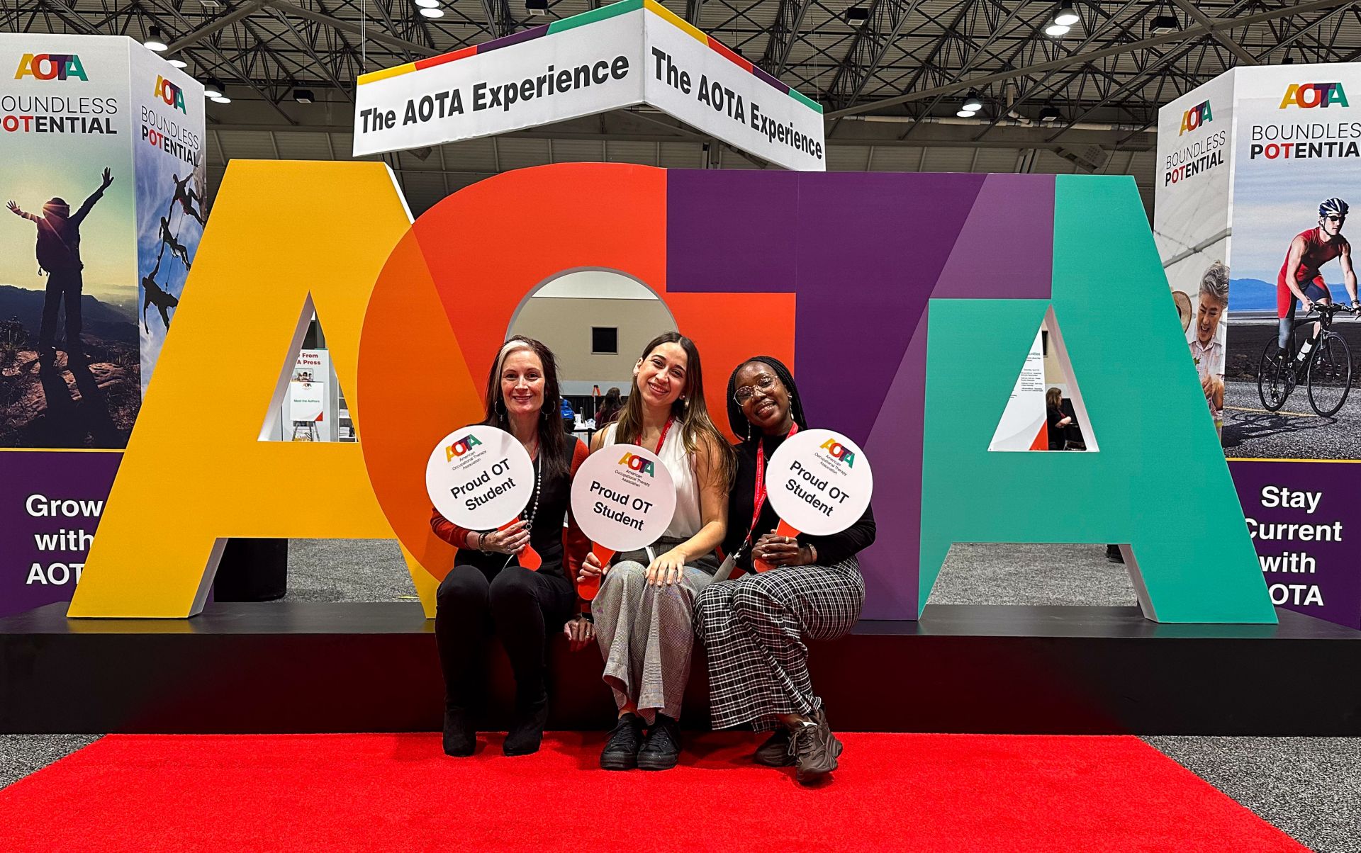Three students smiling in front of large AOTA sign