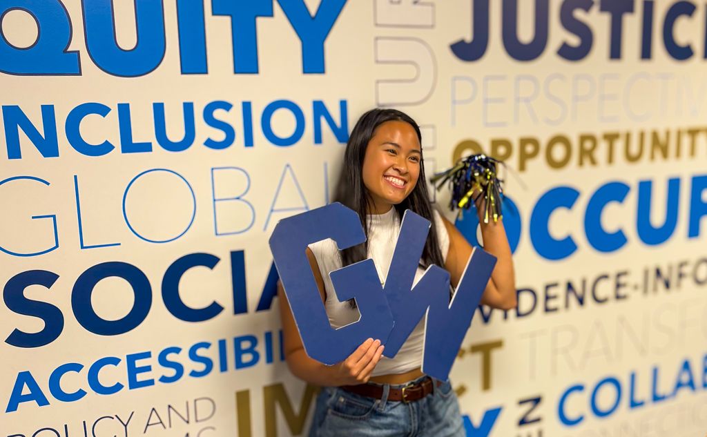 GW OT Student Nicole holding blue GW Letters and pom pom in front of word wall