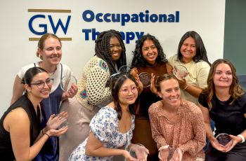 8 Occupational Therapy Students holding circle awards in front of GW OT sign